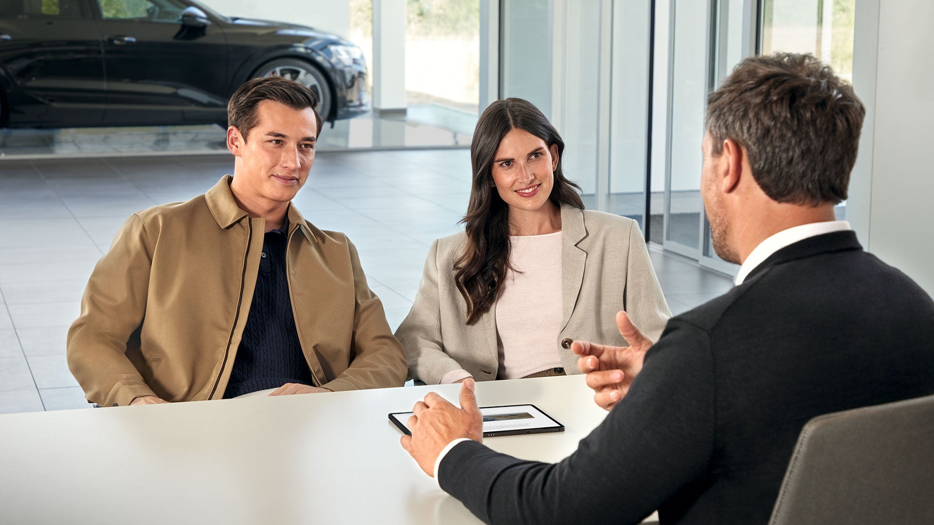 Ein Paar im Gespräch mit einem Audi-Berater am Tisch in einem modernen Autohaus mit Fahrzeug im Hintergrund.