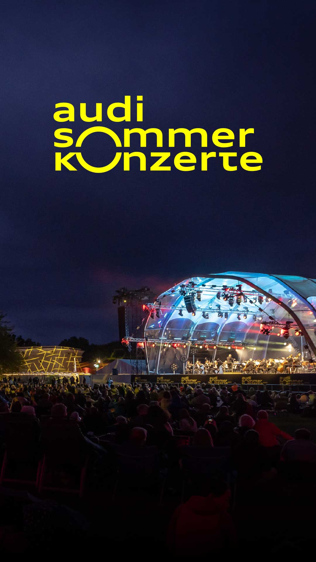 Hell beleuchtete Freiluftbühne am Abend, darüber gelber Schriftzug: Audi Sommerkonzerte.