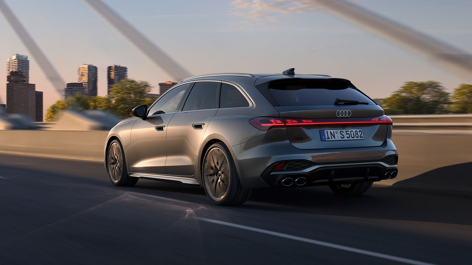 Grauer Audi S5 Avant fährt über eine Brücke mit Stadt-Skyline im Hintergrund bei Sonnenuntergang.