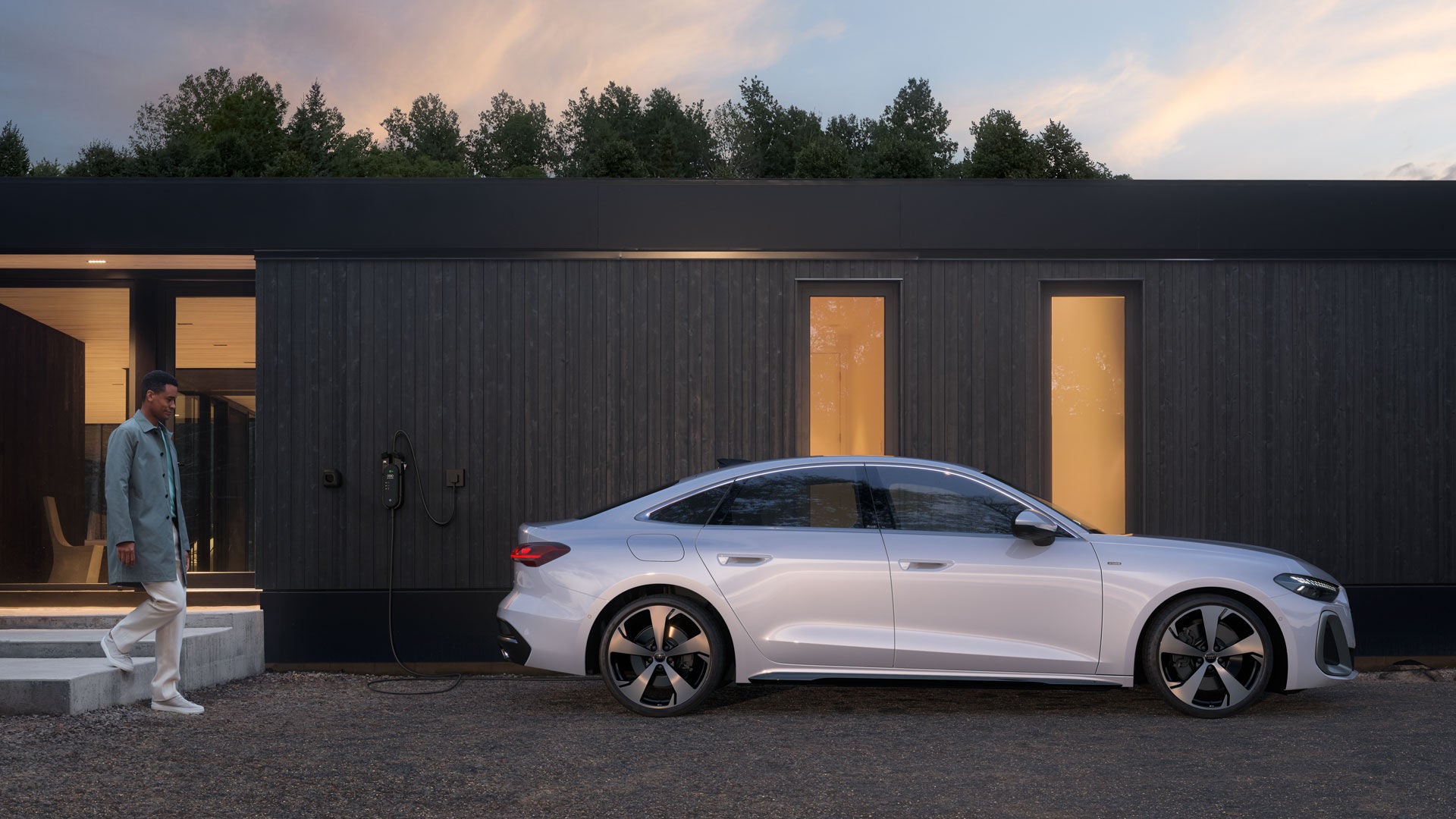 Mann vor modernem Haus neben weißem Audi A5 e-hybrid, der an einer Wallbox-Ladestation angeschlossen ist, bei Abenddämmerung.