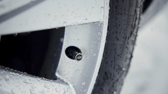 Nahaufnahme eines Autoreifens mit silberner Ventilkappe mit Audi-Ringen, benetzt mit Wassertropfen.