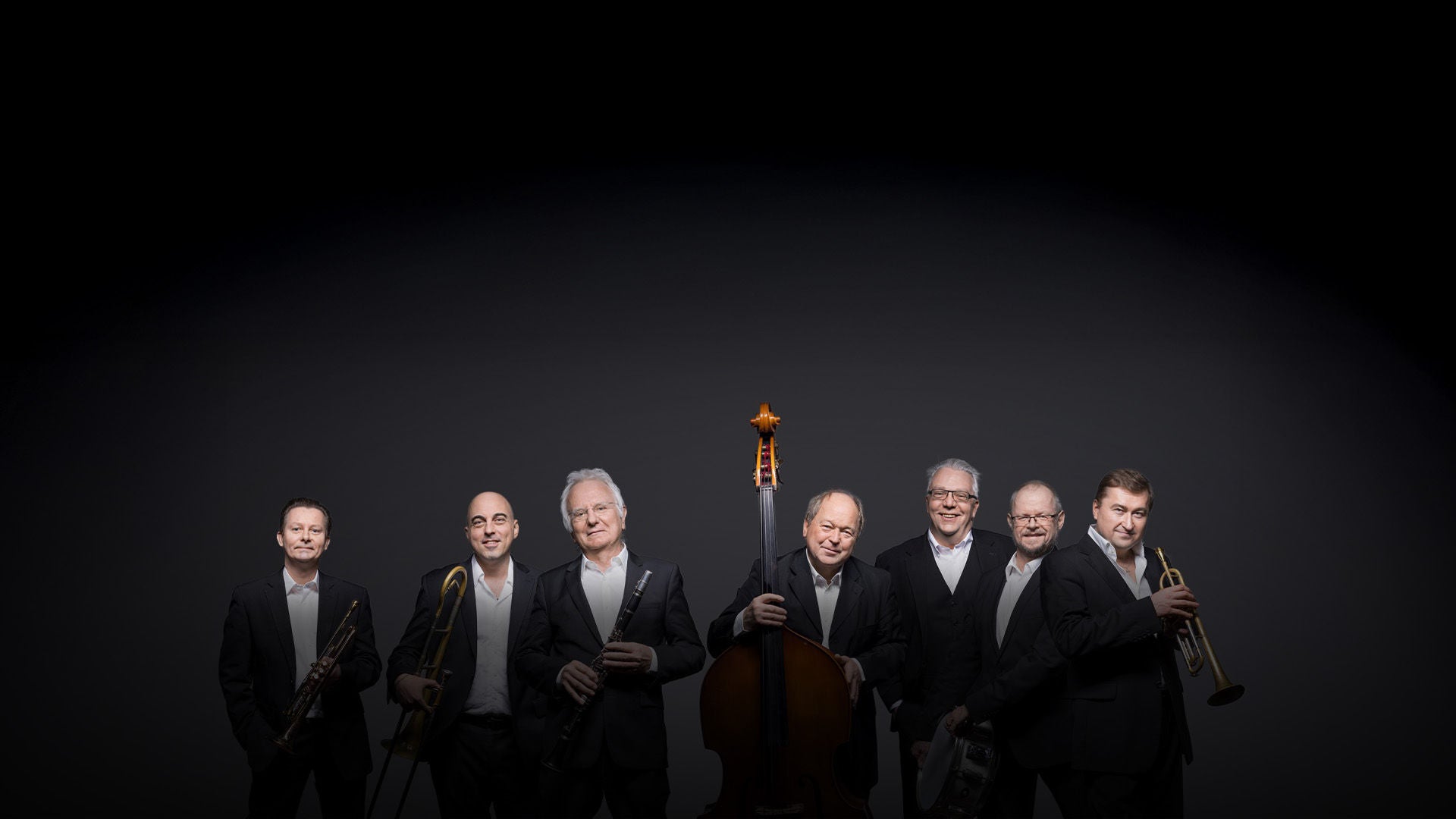 Sieben Musiker in schwarzen Anzügen stehen mit Trompete, Posaune, Klarinette, Kontrabass und Schlagzeug vor grauem Hintergrund.