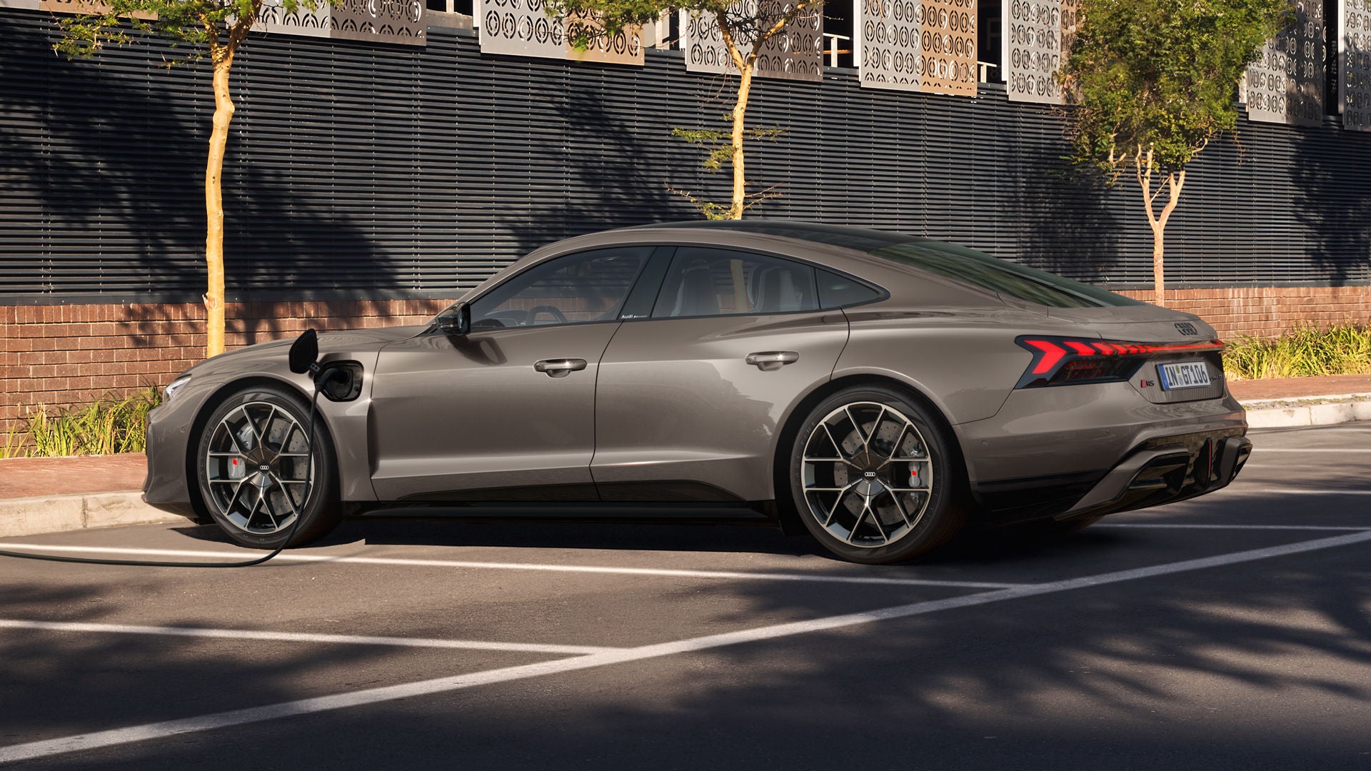Ein lila Audi RS e-tron GT lädt an einer Straßenladestation vor einer modernen Metallfassade mit Bäumen.