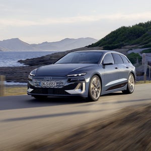 Der S6 Avant e-tron aus schräger Frontansicht, wie er eine Straße an der Küste entlangfährt. Im Hintergrund sind Berge zu sehen.