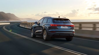 Szenische Fahraufnahme eines Audi Q6 e-tron auf einer Schnellstraße. Rechts von ihm das Wasser, im Hintergrund Berge. Über ihm eine leichte Wolkendecke mit dahinter befindlicher Sonne.