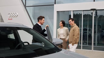 Ein Berater zeigt einem Paar vor einem Audi Autohaus ein geparktes Fahrzeug mit geöffneter Fahrertür