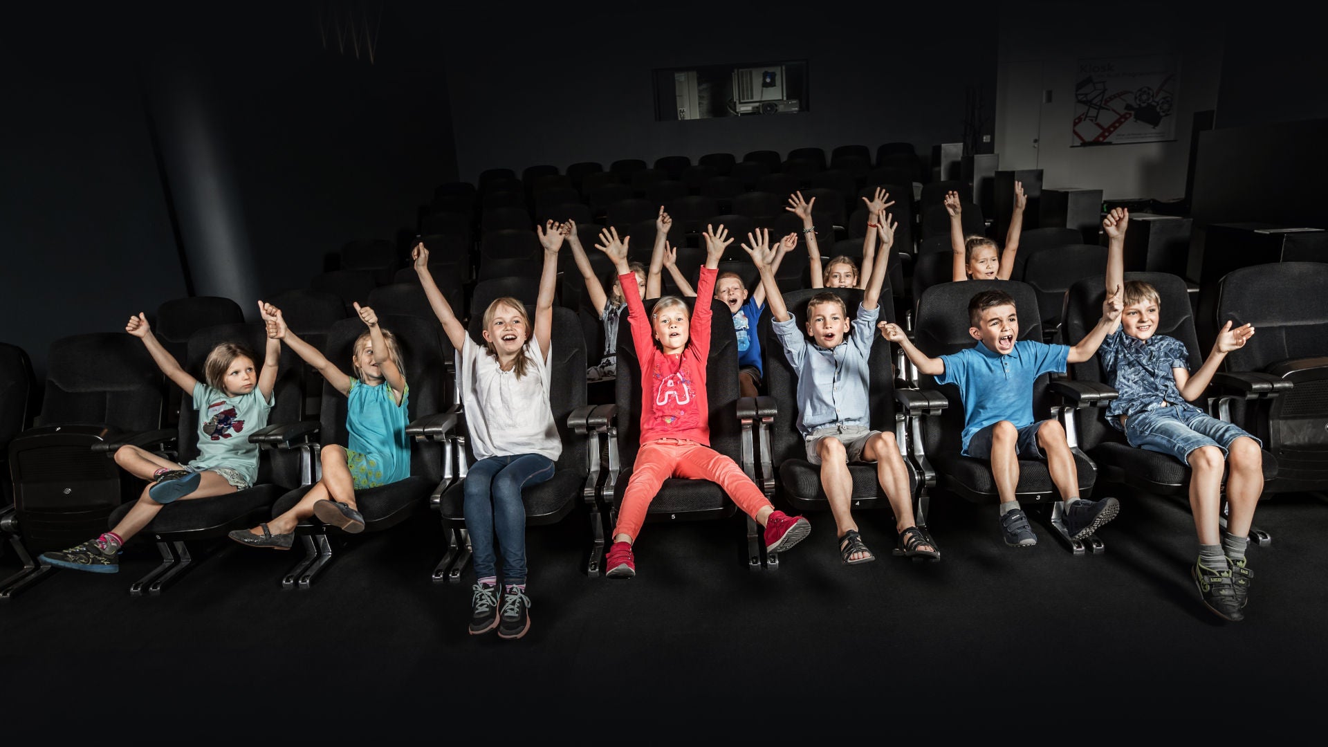 Gruppe begeisterter Kinder in einem dunklen Kinosaal streckt jubelnd die Arme hoch