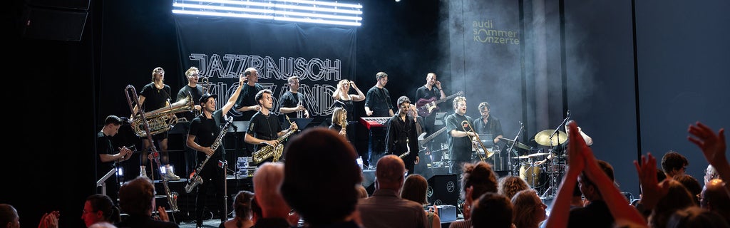 Jazzrausch Bigband performt in schwarzen Outfits energetisch auf der Audi-Sommerkonzerte-Bühne vor jubelndem Publikum.