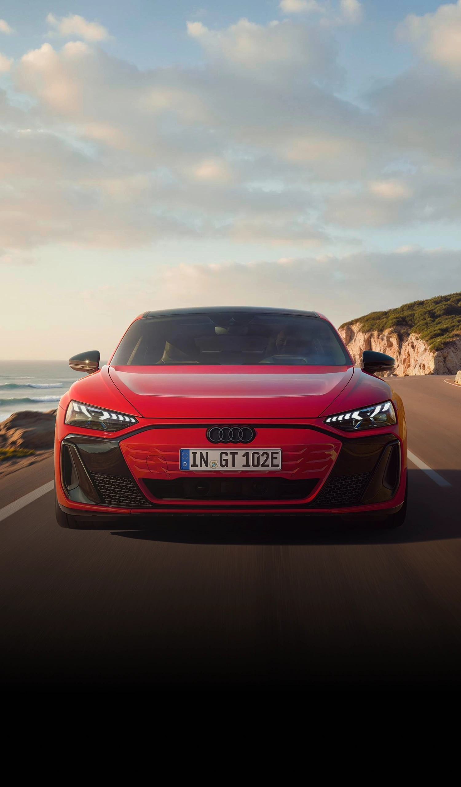Front view of a red Audi e-tron GT driving on a coastal road beside cliffs and the sea.