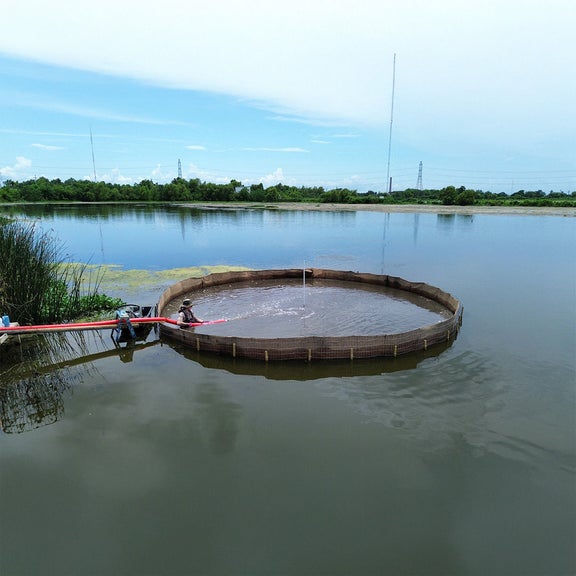 Large round structure in the water with two people and a pump connected to a long hose.