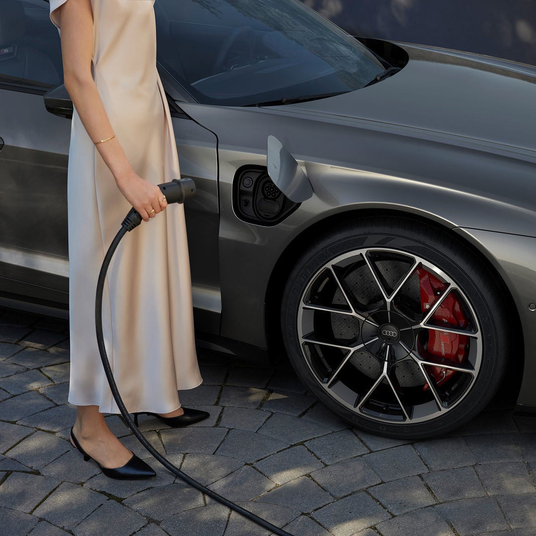 A person in an elegant beige dress charges a sleek grey electric car using a charging cable. The car's wheel features a distinctive, sporty design.