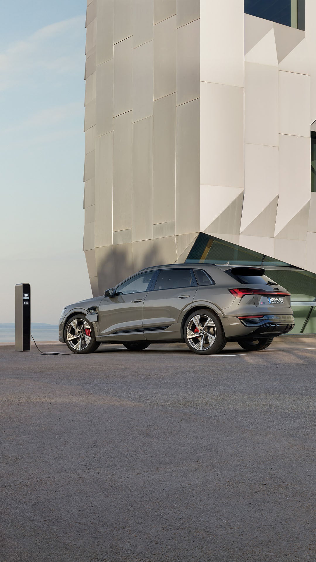 An Audi Q8 e-tron in Chronos Grey Metallic, plugged into a charging station in front of a building.