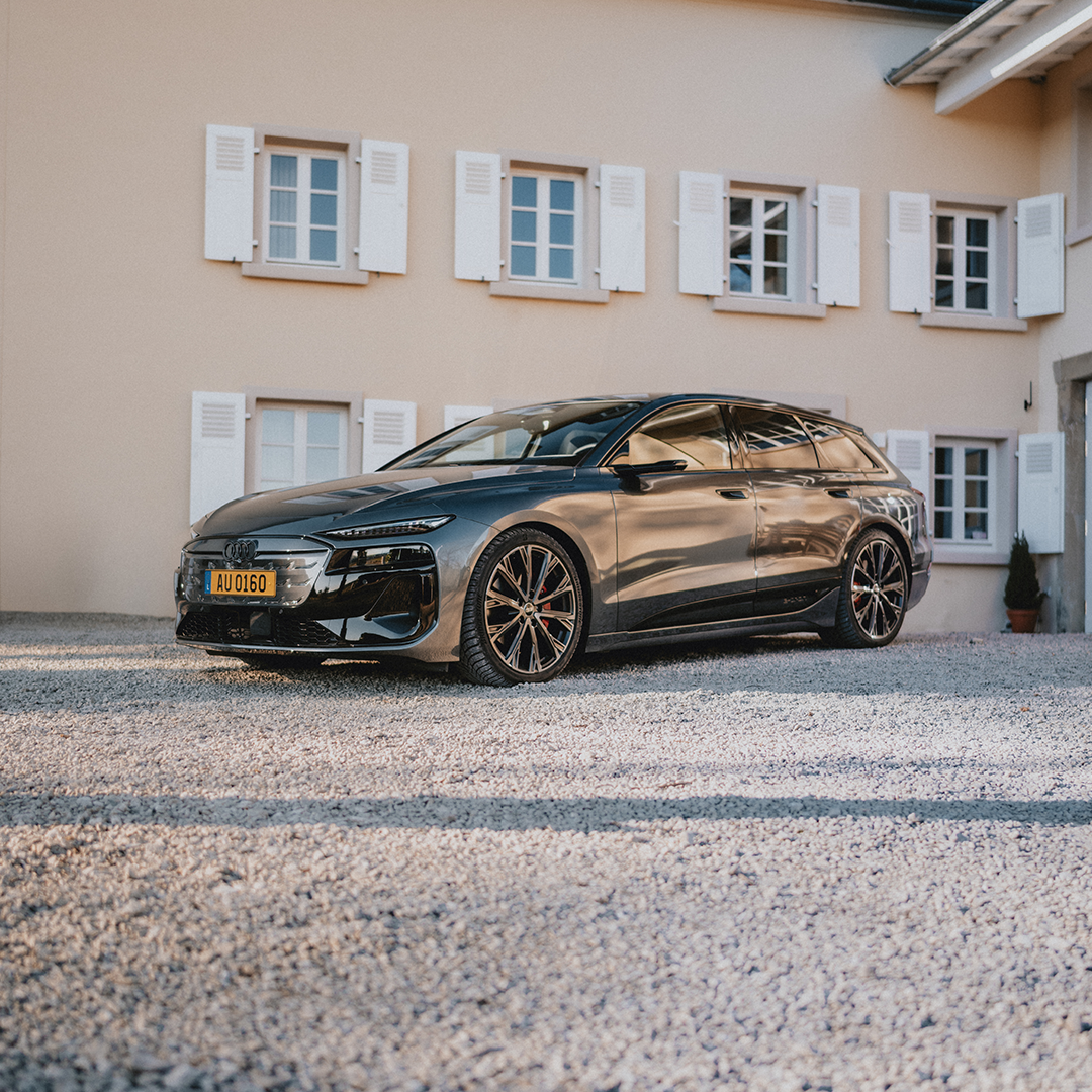 Audi A6 e-tron steht auf einem Schotterhof vor einem beigen Haus mit weißen Fensterläden.