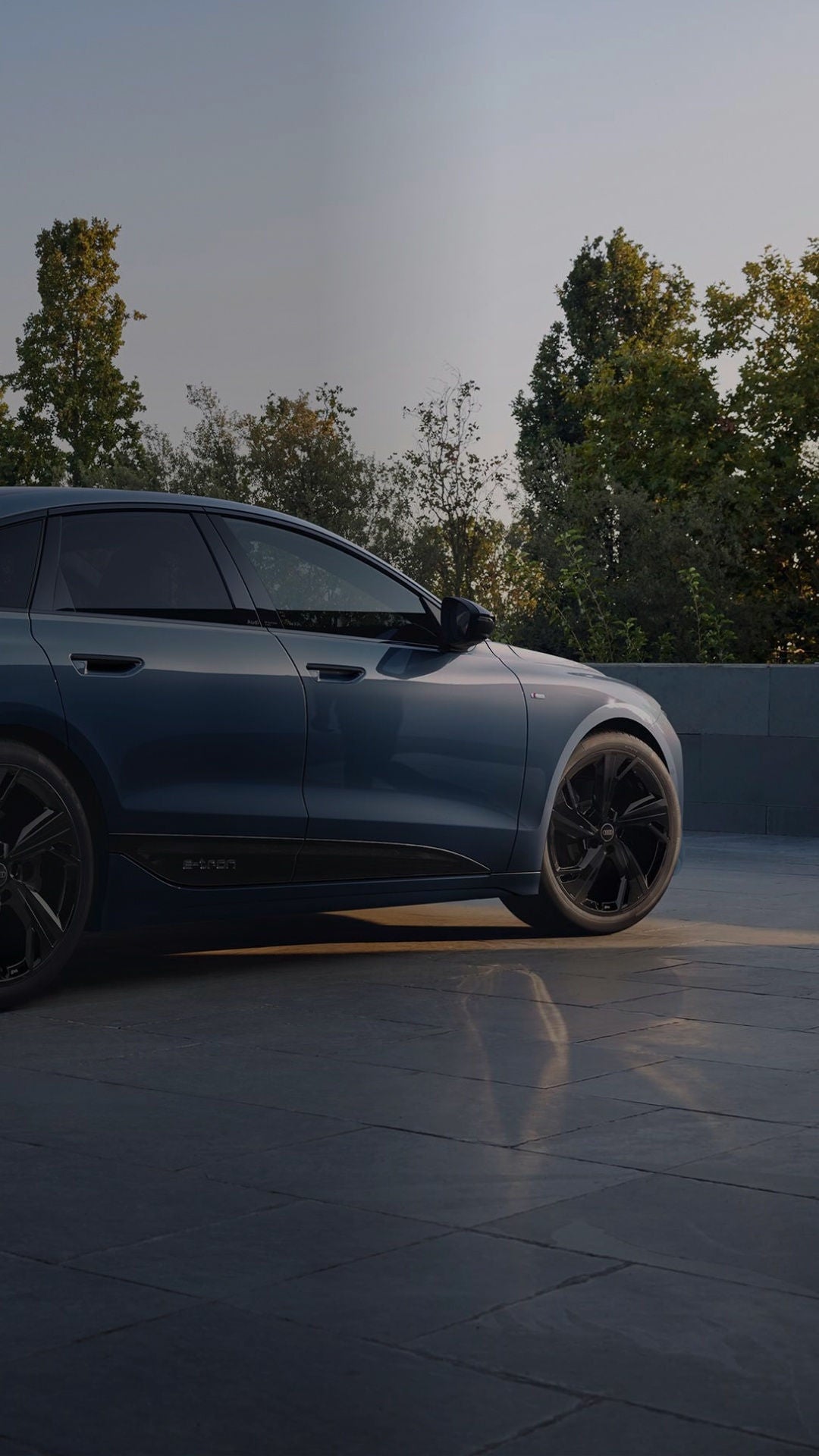 An Audi is parked on a spacious, modern stone driveway. A man in a green sweater walks nearby. Lush trees are in the background.