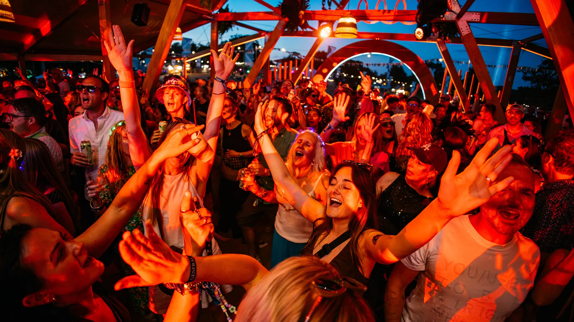A joyful nighttime crowd wearing light-up crowns and glittery face decorations sings and dances together at the festival. A joyful nighttime crowd wearing light-up crowns and glittery face decorations sings and dances together at the festival.
