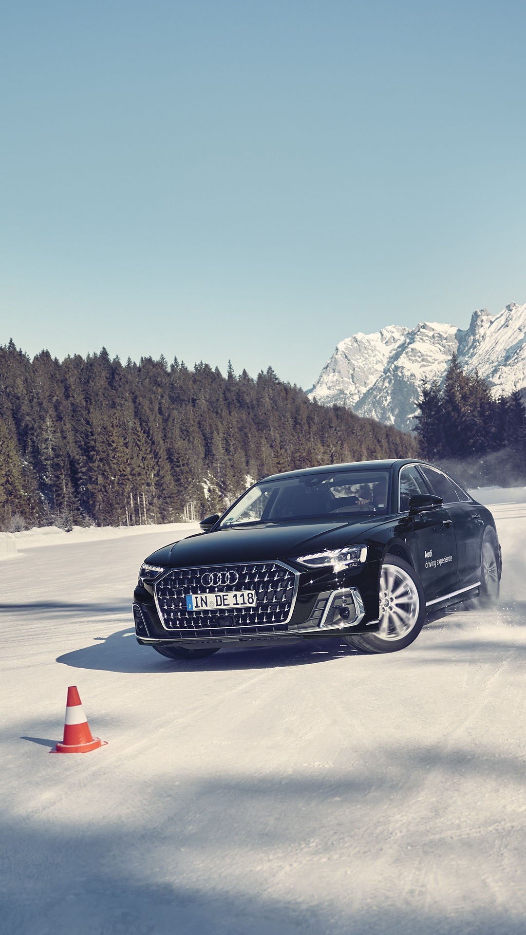 Schwarzer Audi A8 50 TDI quattro fährt im Schnee durch Parcour, im Hintergrund blauer Himmel, Wälder und Berge.