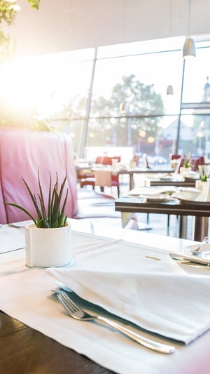 Set tables at the Nuvolari restaurant, dressed in white with a plant; the seating is red; in the background, a glass front offering a view outside in daylight.