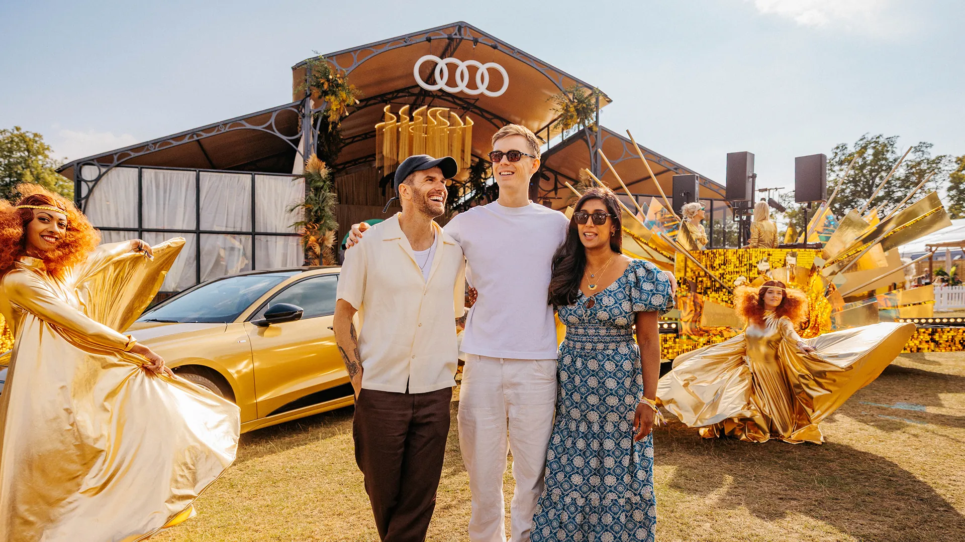 Three people laugh together arm-in-arm in front of an Audi-branded festival pavilion, with a gold electric Audi and performers in shimmering gold costumes nearby. Three people laugh together arm-in-arm in front of an Audi-branded festival pavilion, with a gold electric Audi and performers in shimmering gold costumes nearby.