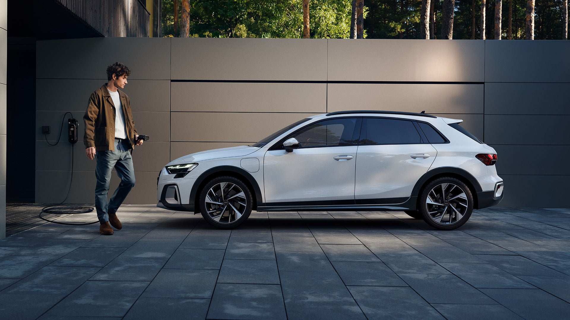 Ein Mann mit Ladekabel steht vor einem weißen Audi A3 allstreet TFSI e, der vor einer modernen Wand geparkt ist.