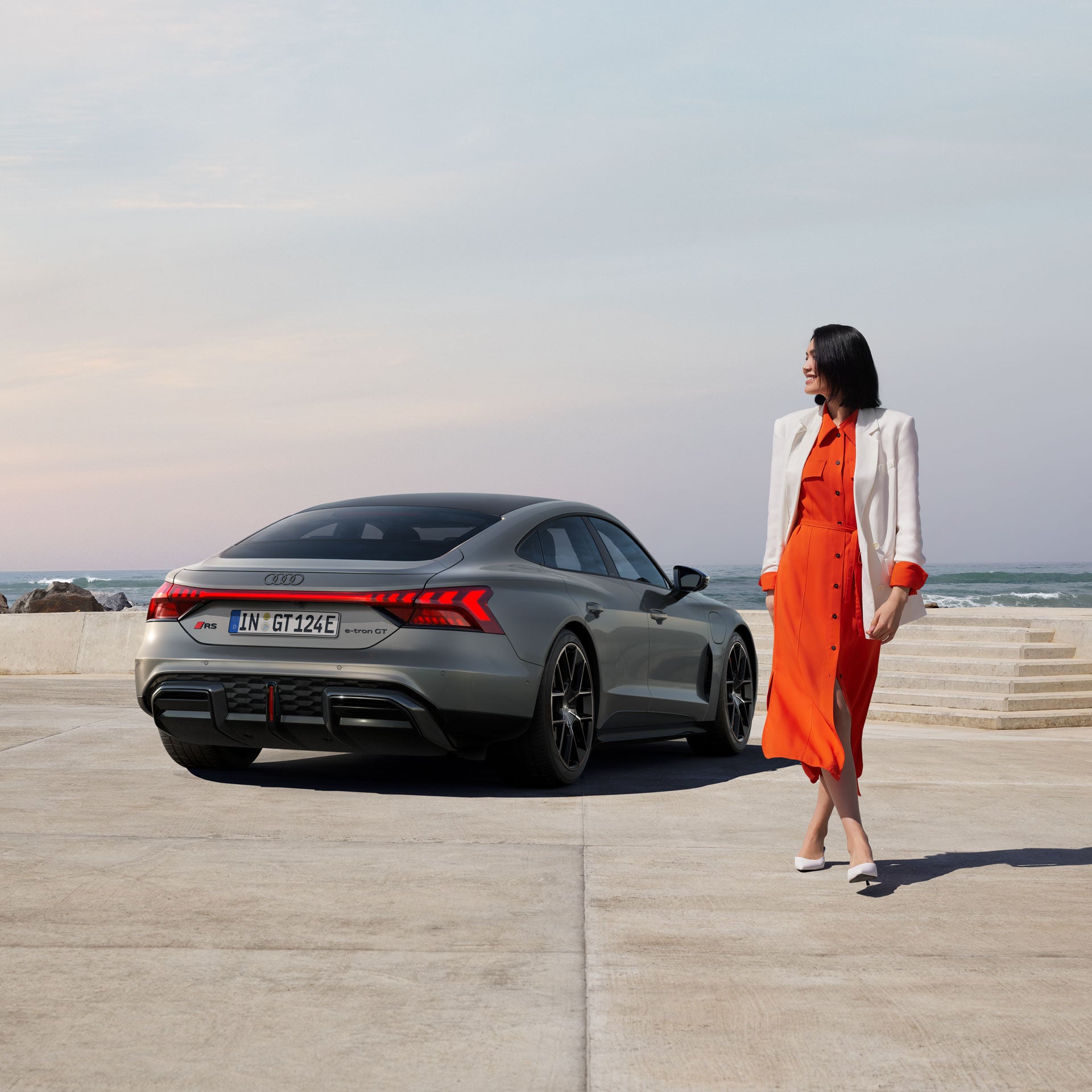 Frau in rotem Kleid und weißem Blazer geht neben einem grauen Audi RS e-tron GT am Meer entlang.