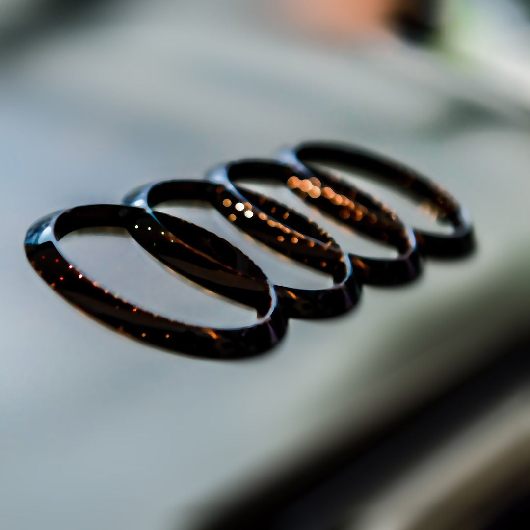 Nahaufnahme des Audi-Logos mit vier verchromten Ringen auf unscharfem, grauem Hintergrund.