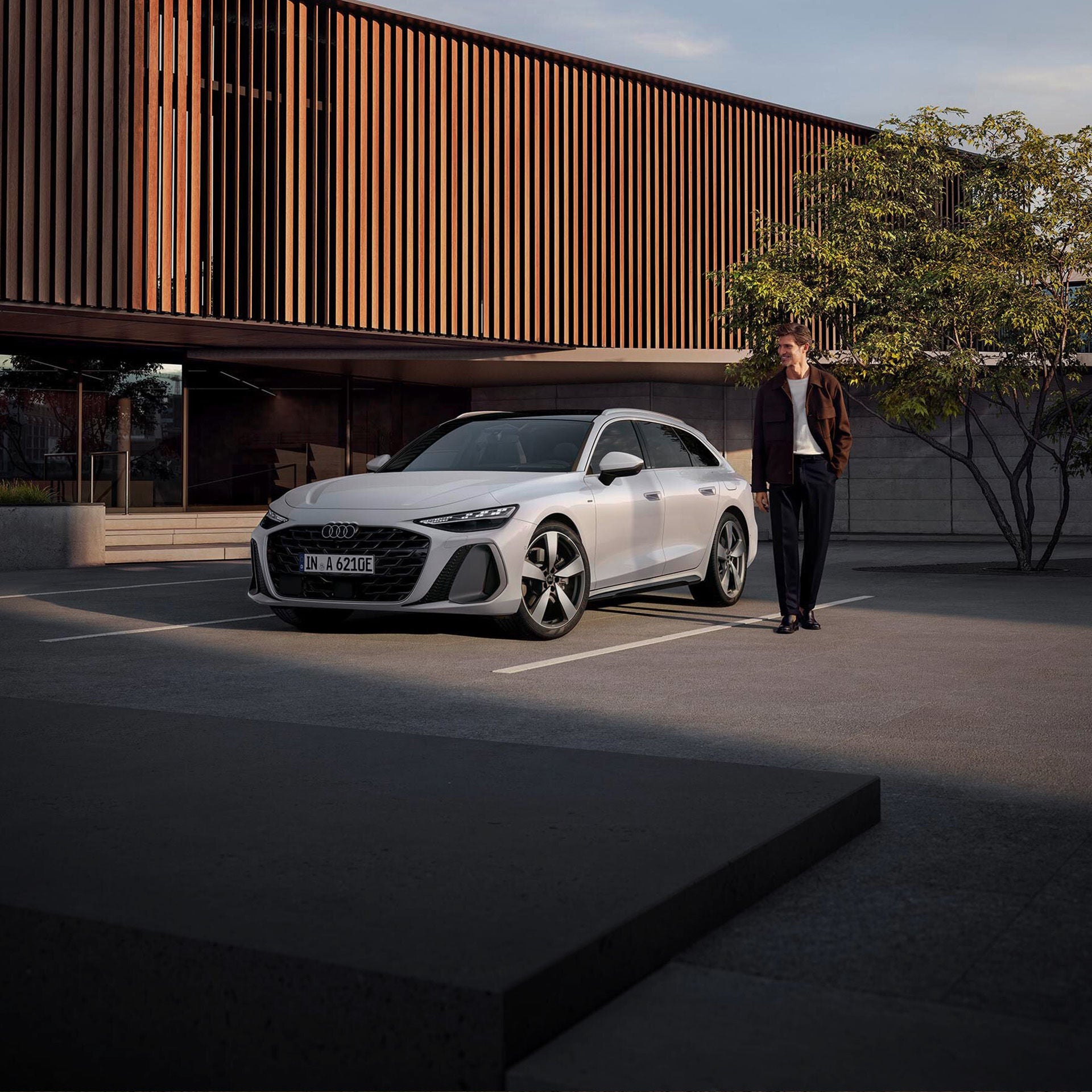 Ein junger Mann steht neben einem weißen Audi A6 e-hybrid auf einem Parkdeck vor moderner Architektur.