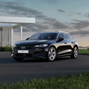 Une A5 Avant noire est garée devant une maison moderne aux murs vitrés, à la lisière de la nature.