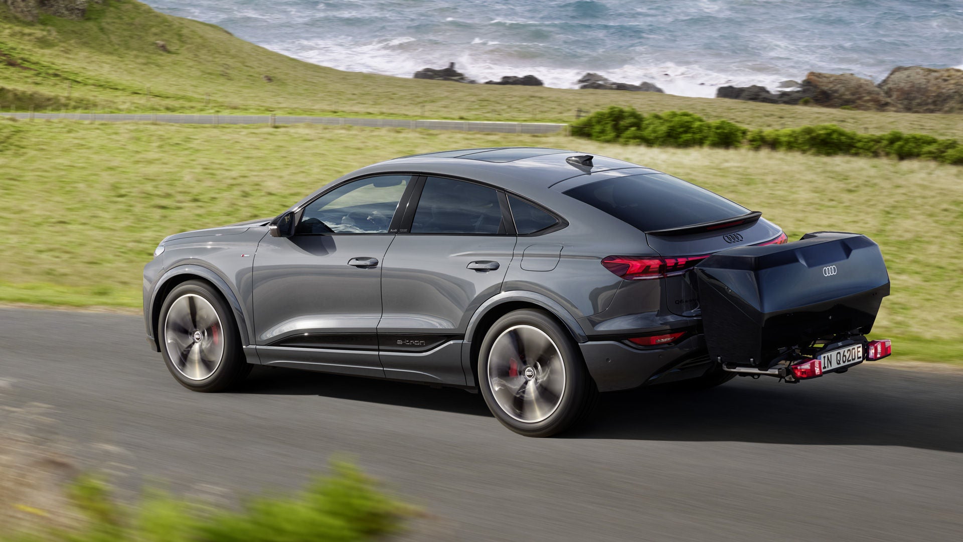 The Q6 Sportback e-tron with rear box drives along a country road surrounded by nature.
