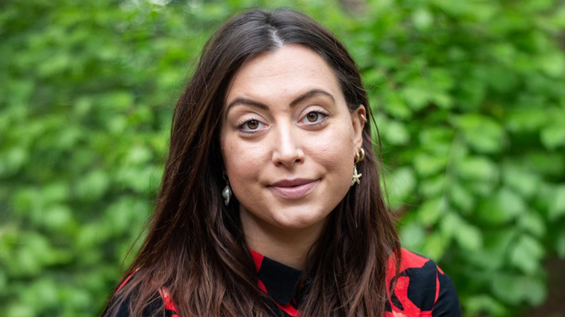 Porträt einer Frau mit langen braunen Haaren, rotem und schwarzem Oberteil, vor grünem, unscharfem Hintergrund.