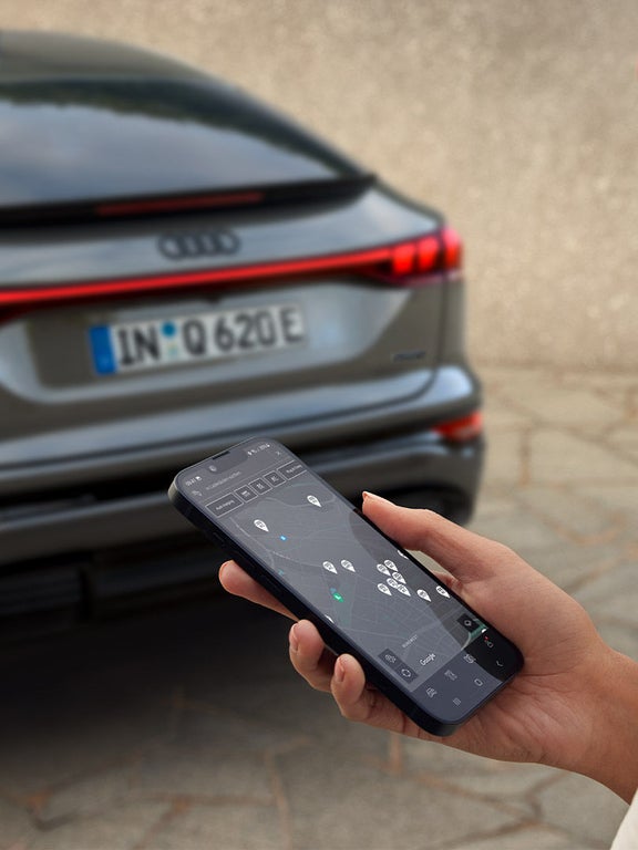 Person hält Smartphone mit myAudi App vor dem Heck eines grauen Audi SQ6 Sportback e-tron mit sichtbar leuchtendem Rücklicht.