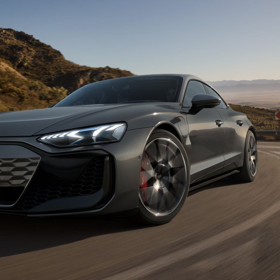 Ein grauer Audi RS e-tron GT fährt auf einer kurvigen Bergstraße bei Sonnenlicht, mit leuchtenden LED-Scheinwerfern.