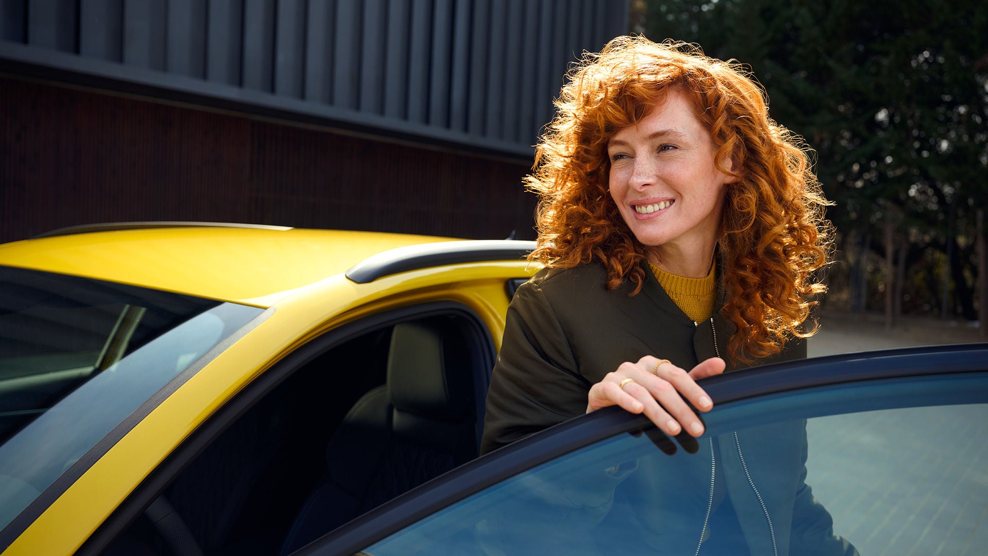 Lächelnde Frau mit roten Locken steigt bei Sonnenschein aus einem gelben Audi A3, Tür geöffnet, moderne Kulisse im Hintergrund.