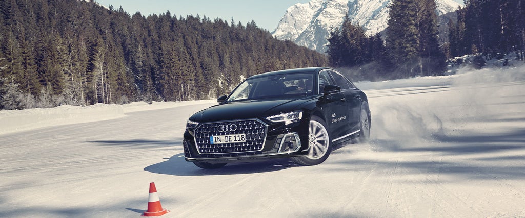 Schwarzer Audi A8 50 TDI quattro fährt im Schnee durch Parcour, im Hintergrund blauer Himmel, Wälder und Berge.