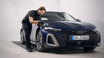 Mann poliert die Motorhaube eines blauen Audi mit Poliermaschine in heller Werkstattumgebung.