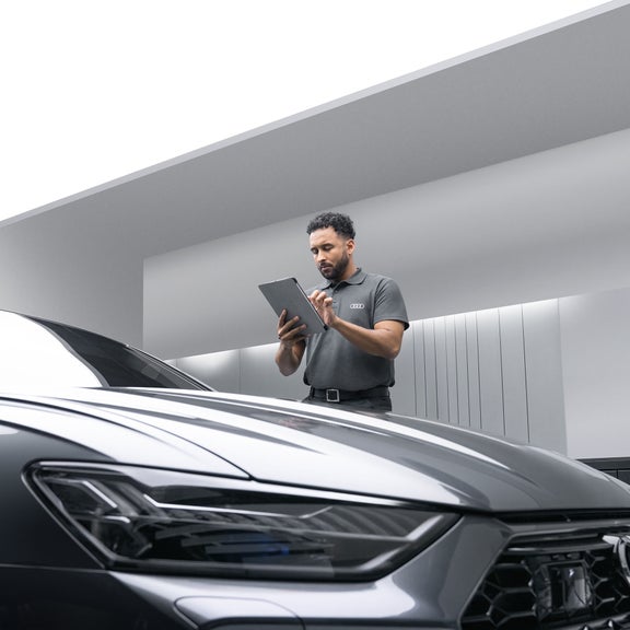 A male Audi Service representative using a tablet computer while standing next to a dark grey Audi.