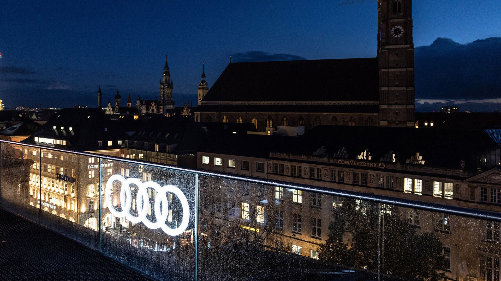 Blick über München bei Nacht mit beleuchtetem Audi-Logo im Vordergrund und Frauenkirche im Hintergrund.