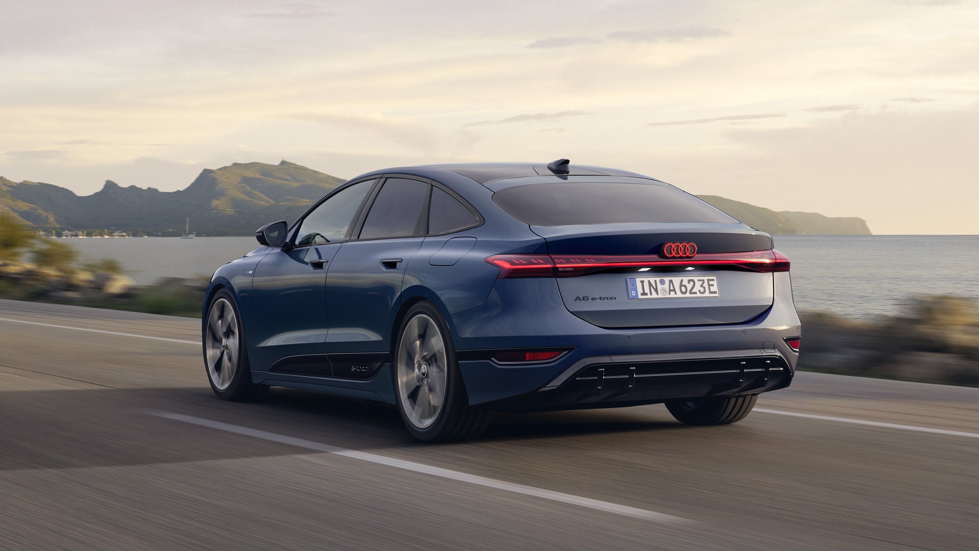 Der A6 Sportback e-tron aus schräger Heckansicht fährt eine Straße parallel zum Ufer entlang. Im Hintergrund sind Berge zu erkennen.&nbsp;