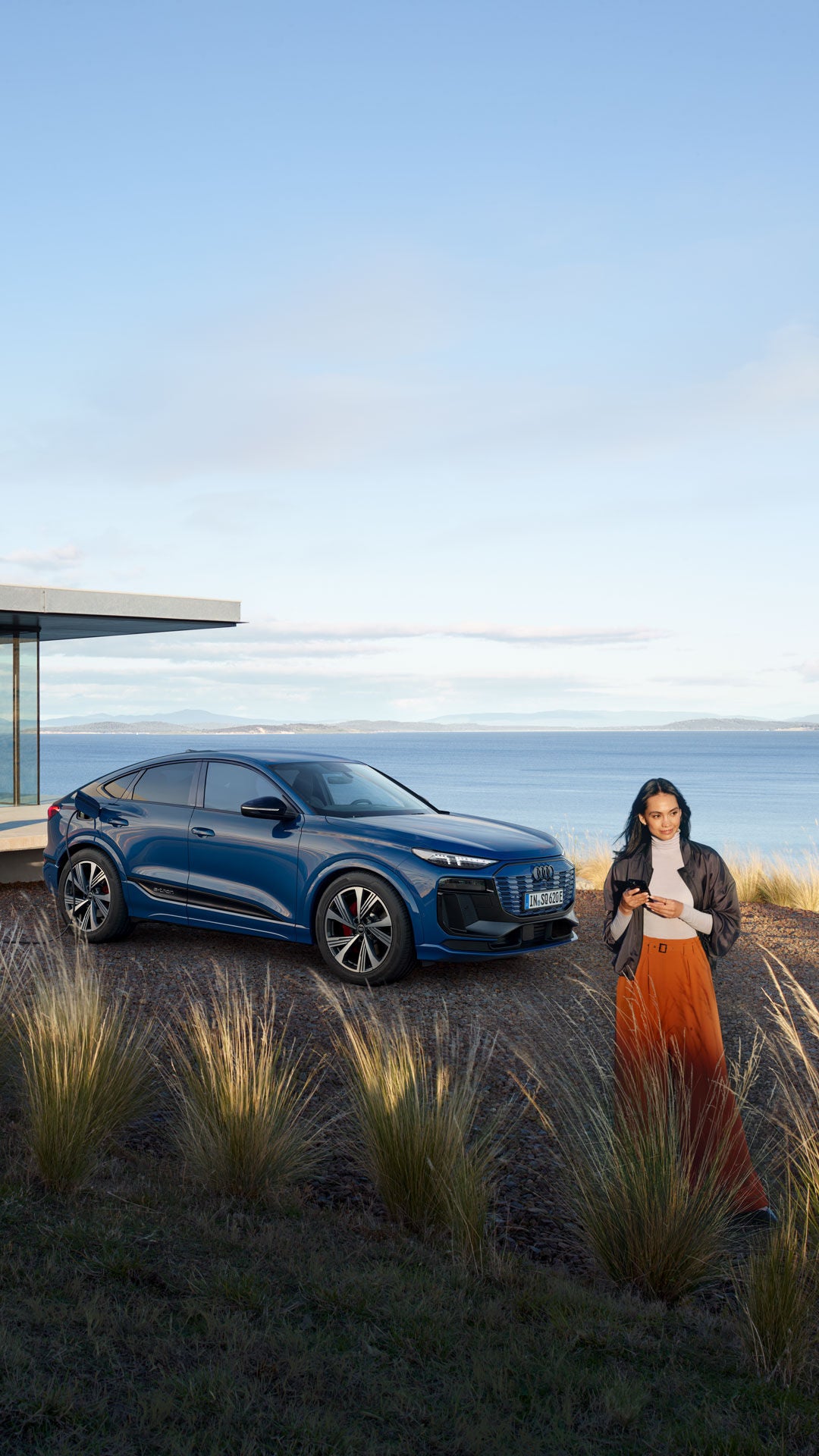 Frau steht vor einem blauen Audi SQ6 Sportback, der an einer Ladestation nahe eines modernen Hauses am Wasser geladen wird.
