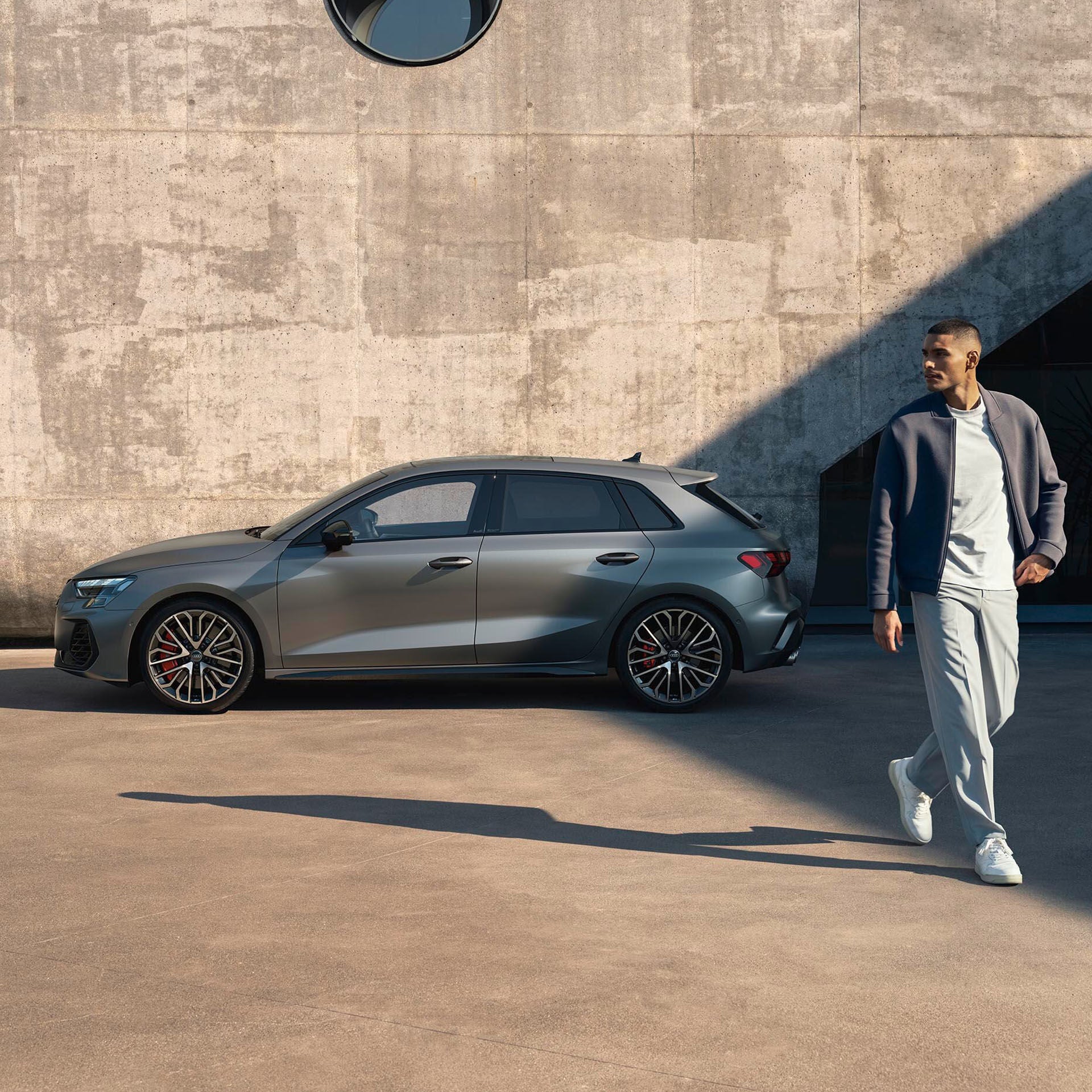 Mann geht an einem grauen Audi S3 Sportback vorbei, im Hintergrund eine moderne Betonfassade mit rundem Fenster.