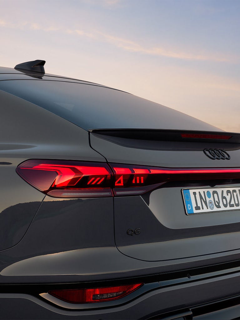 Nahaufnahme des Hecks eines grauen Audi Q6 Sportback e-tron mit beleuchteten Rückleuchten bei Sonnenuntergang.
