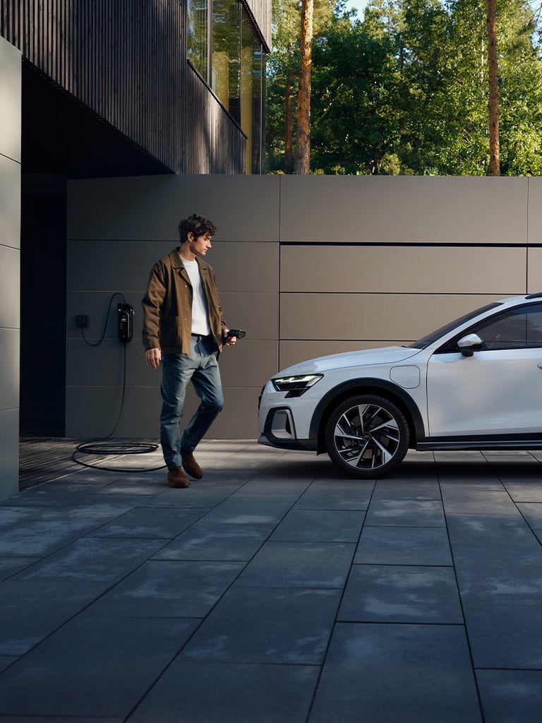 Mann mit Ladekabel steht neben weißem Audi A3 allstreet TFSI e vor moderner Hauswand.