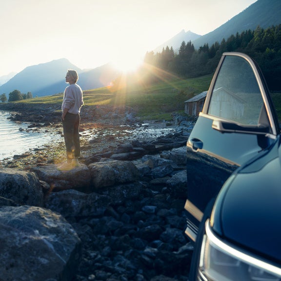 Landskap med berg och hav, en person vid stranden en bil med öppen dörr i förgrunden.