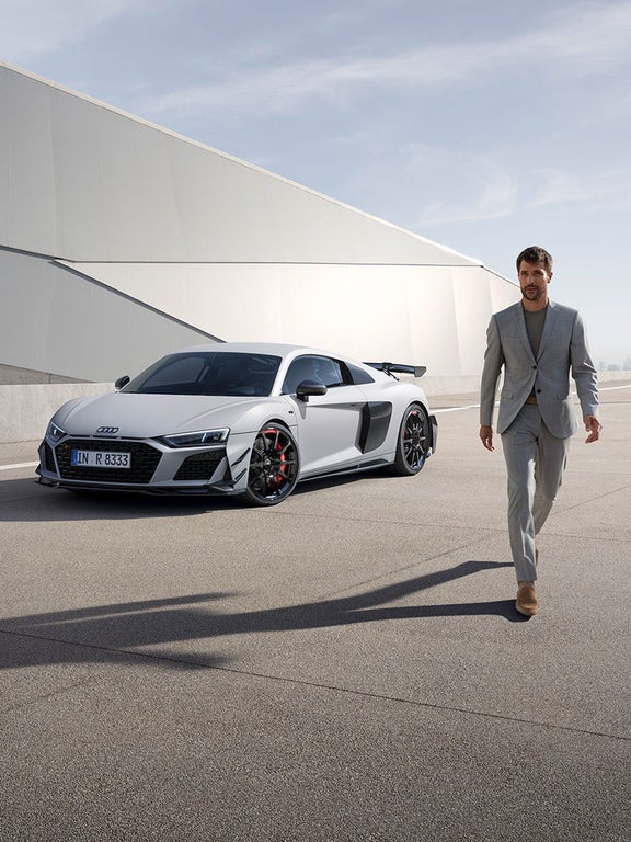 A man walking towards the camera. A white Audi R8 GT is behind him.