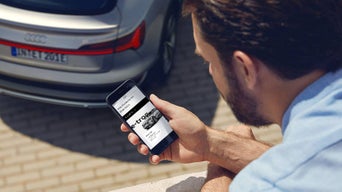 Ein Mann schaut auf sein Handy im Hintergrund das Eck eines silbernen Audis
