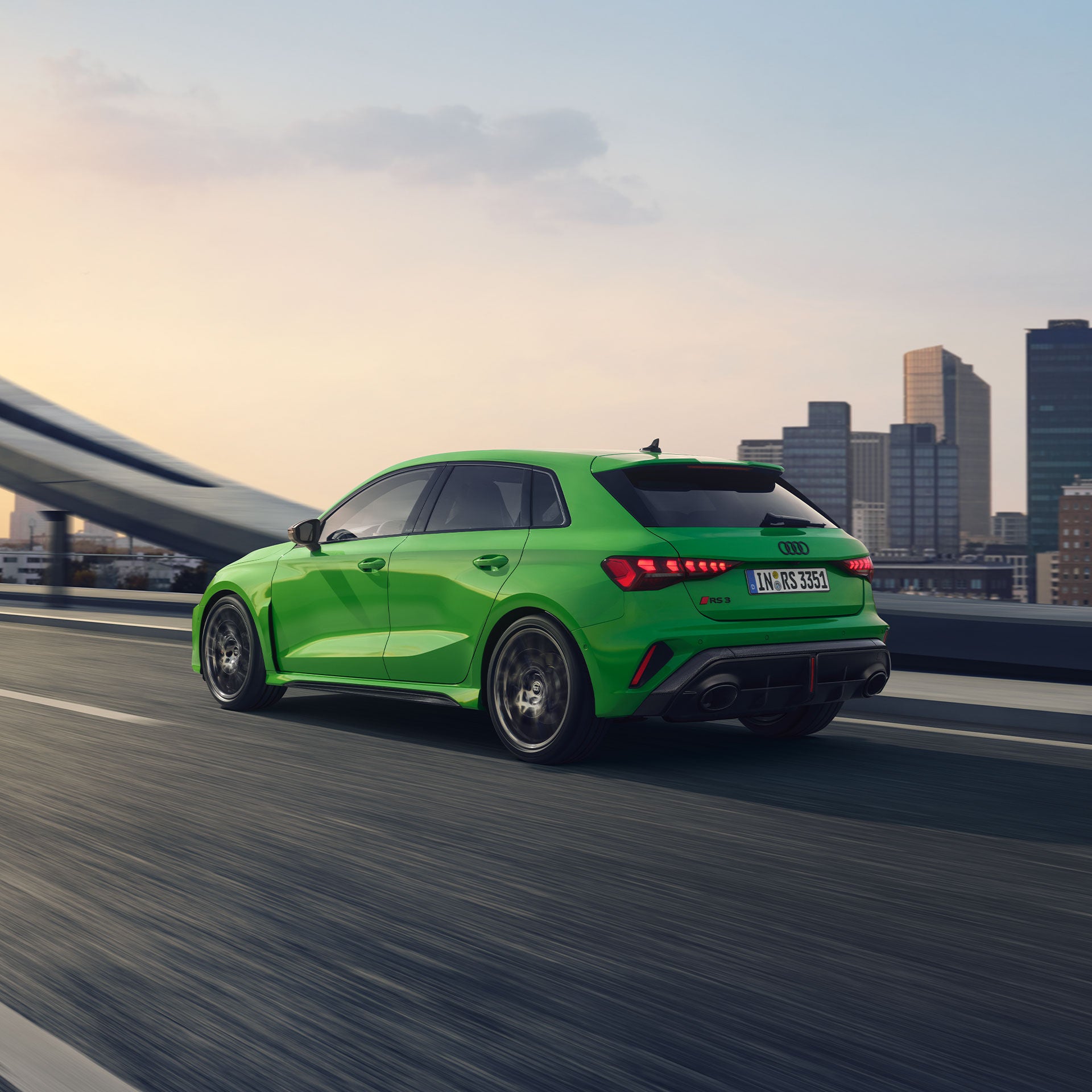 Grüner Audi RS 3 Sportback fährt auf einer Brücke vor städtischer Skyline, Rückansicht.