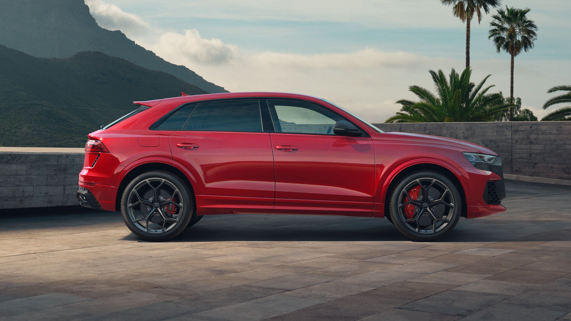 De RS Q8 SUV Performance geparkeerd vanaf de zijkant.  De muur erachter markeert de eigendomsgrens, met palmbomen aan de rechterkant en torenhoge bergen aan de linkerkant.