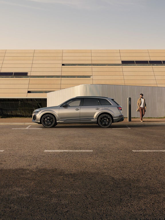 Ein silberner Audi Q7 SUV TFSI e an Ladesäule, Mann im beigen Mantel nähert sich, moderne helle Gebäude im Hintergrund.