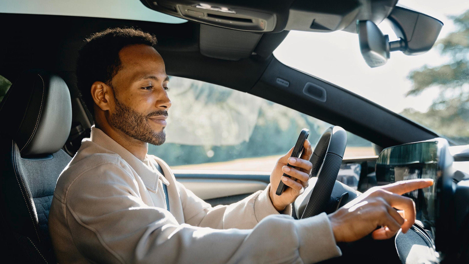 Ein Mann sitzt in einem Auto, hält ein Smartphone in der einen Hand und bedient mit der anderen das Touchscreen-Display des Armaturenbretts.