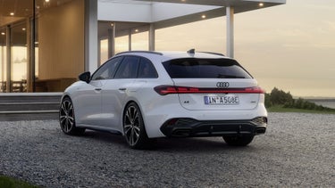 The A5 Avant e-hybrid is shown from a slanted rear perspective on a gravel surface in front of a modern residential building.