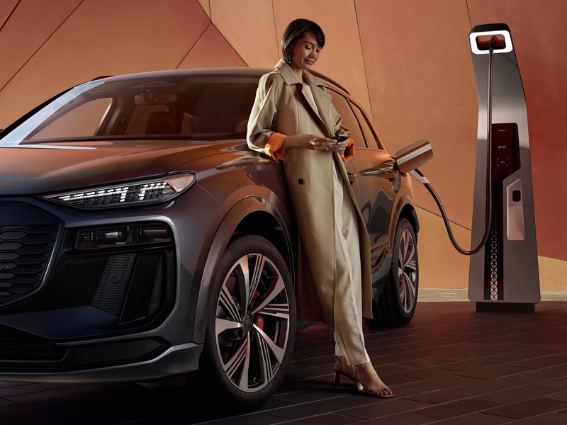 De SQ6 SUV e-tron staat geparkeerd voor een oranje gebouwgevel en is aangesloten op een laadstation. Een jonge vrouw leunt tegen de zijkant van de auto en kijkt naar haar mobiele telefoon.